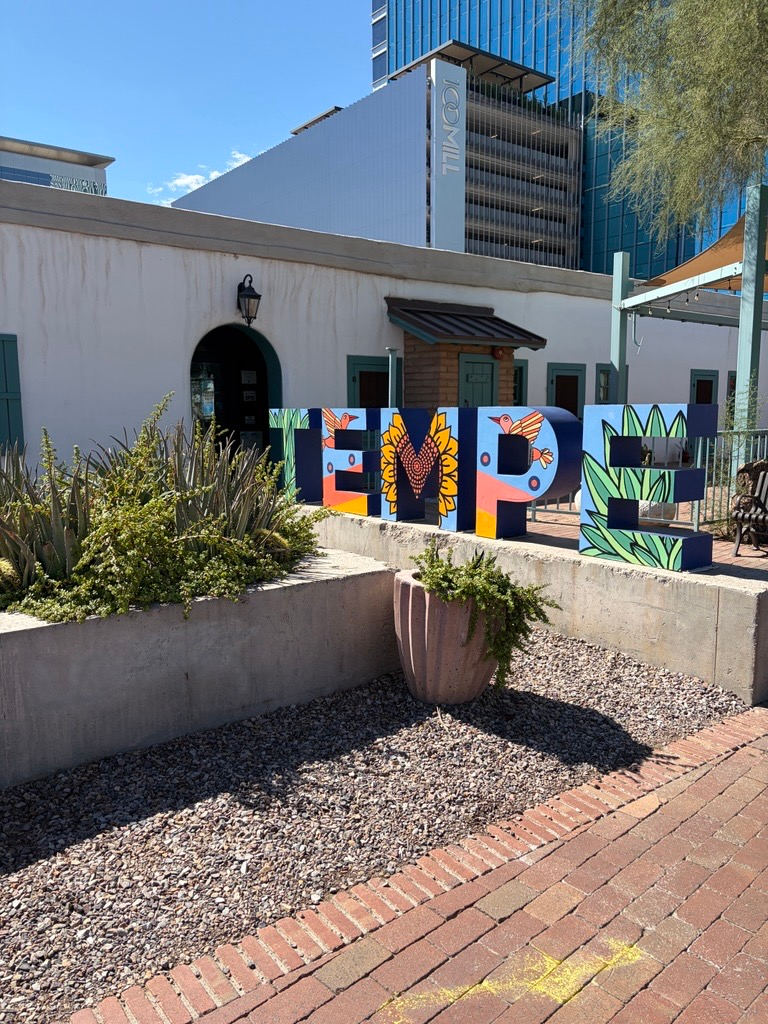 Tempe, Arizona sign on a sunny day. The Tempe letters are blue and decorated with painted elements from the desert: cacti, birds, sunflowers, and a sunset.